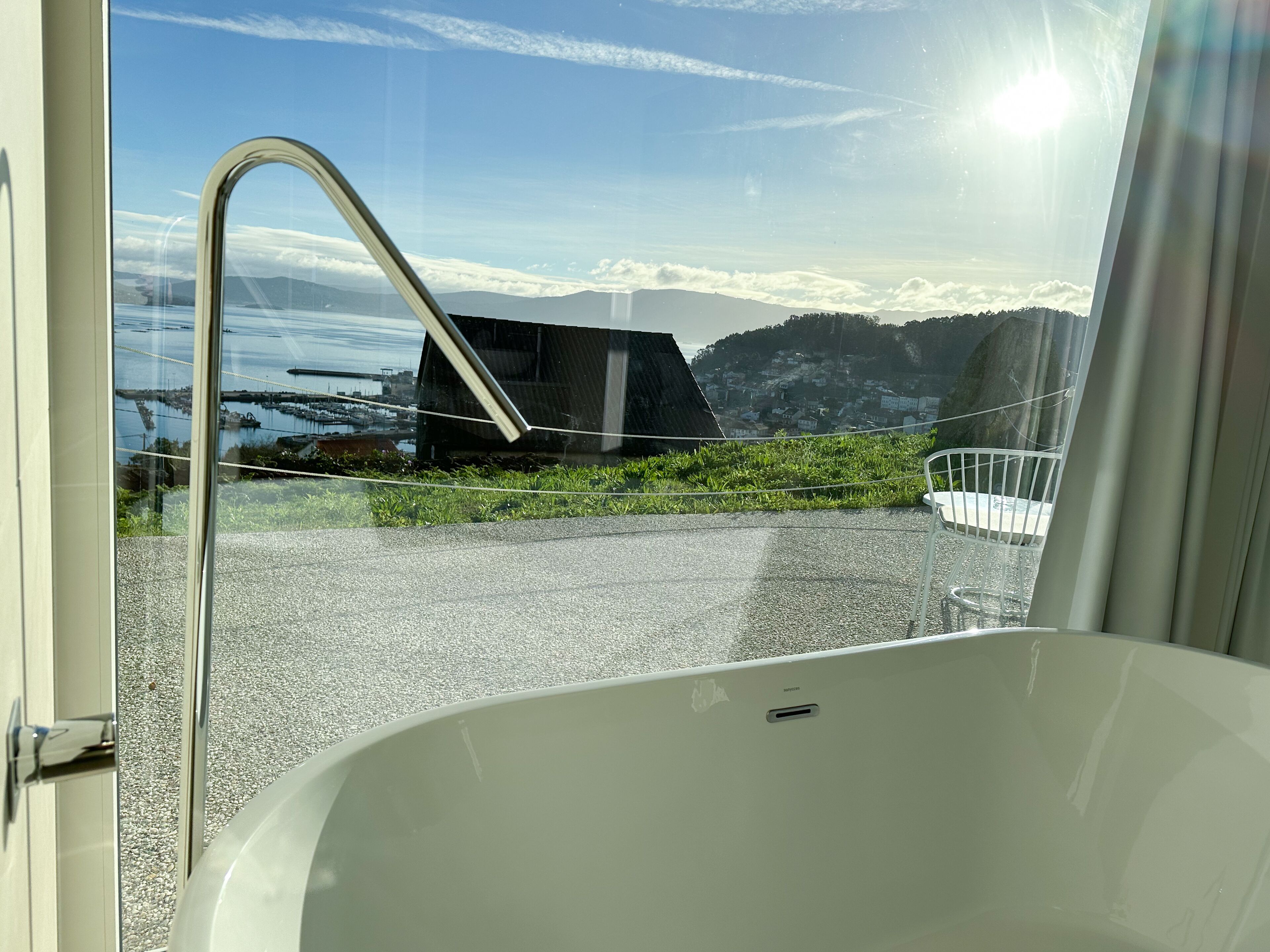 A modern bathroom with a large bathtub, a glass door leading to a balcony with a scenic view of a coastal area, and a metal railing.