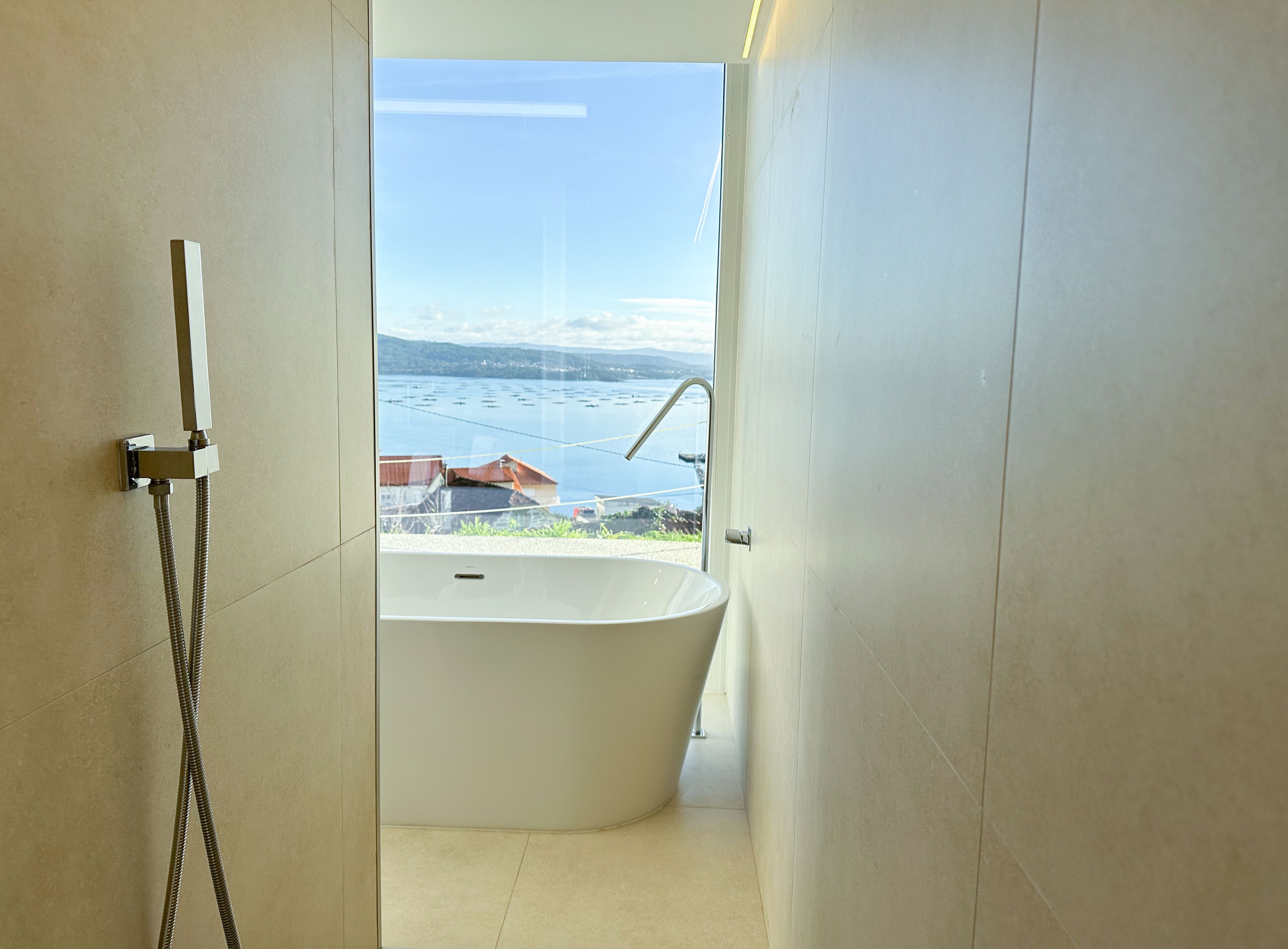 A modern bathroom with a freestanding bathtub, a showerhead, and a view of a lake and mountains.