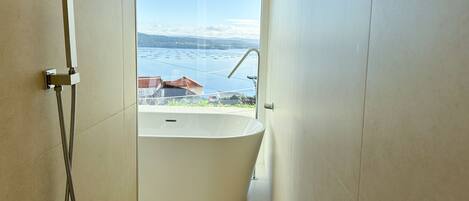 A modern bathroom with a freestanding bathtub, a showerhead, and a view of a lake and mountains.