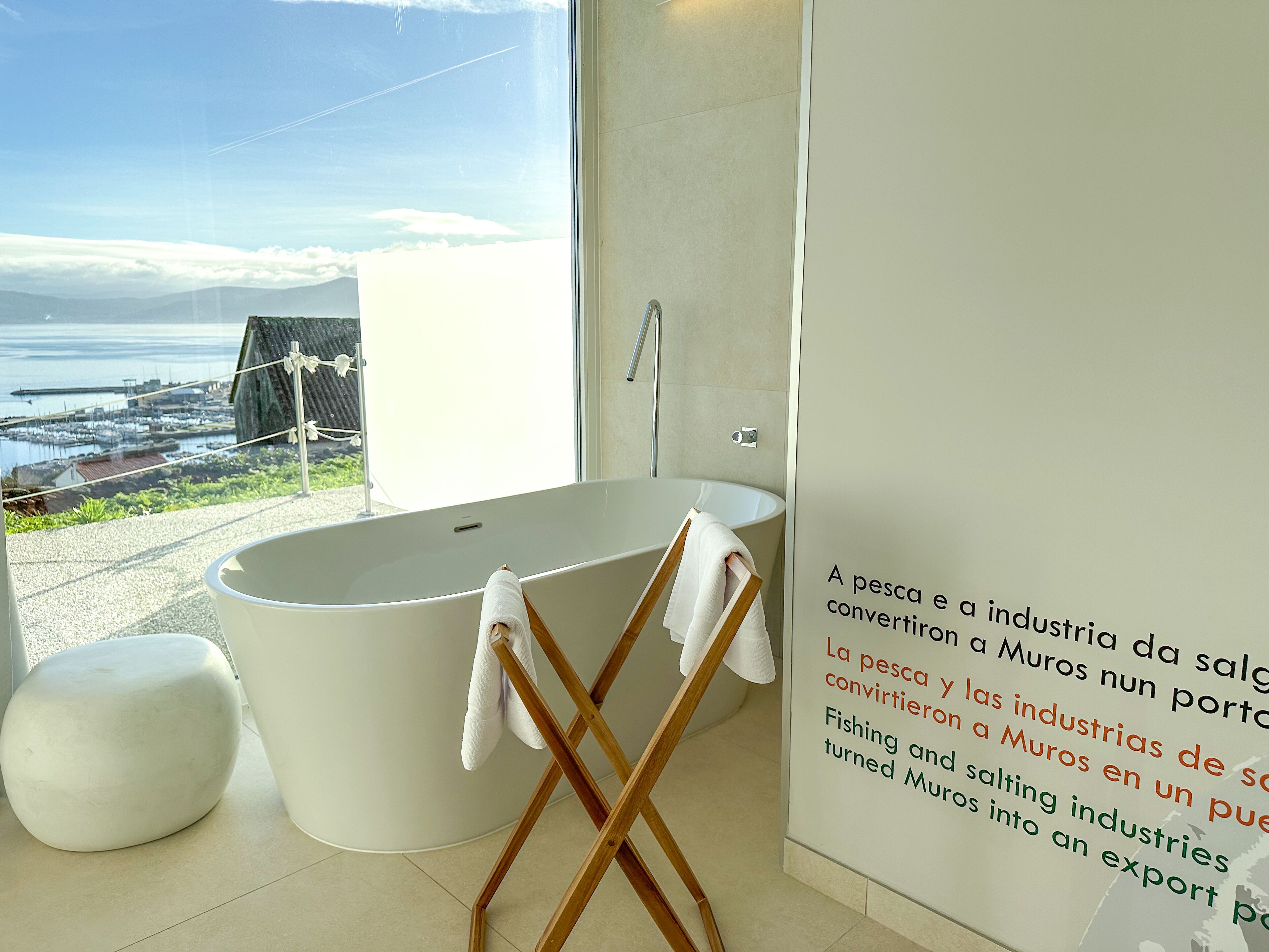 Un baño moderno con una bañera grande, una silla de madera y vistas al mar y el puerto deportivo.