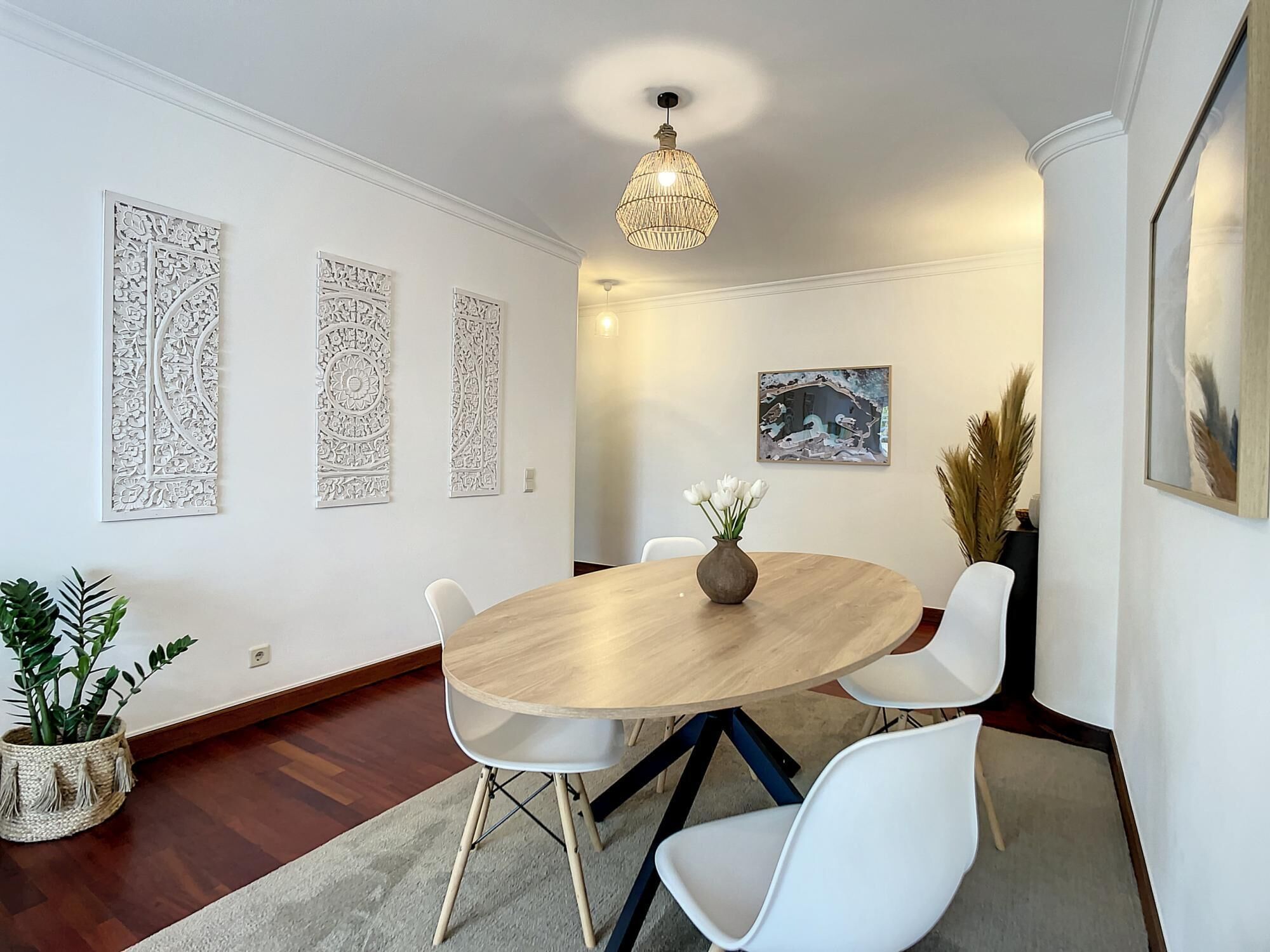 A dining area with a round wooden table, white chairs, a vase with flowers, and decorative wall art.