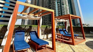 Zona de estar en la azotea con pérgolas de madera y sillones azules, con vistas a edificios altos.