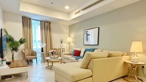 Una sala moderna con un sofá beige, una mesa de centro redonda y una mesita de noche con una lámpara. Hay un ventanal con cortinas, un cuadro en la pared y una planta en maceta.