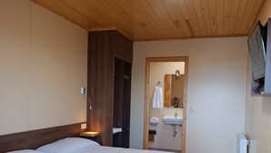 A hotel room with a wooden ceiling, a large bed with white bedding, a bedside table, a wardrobe, a bathroom with a sink and mirror, and a television mounted on the wall.