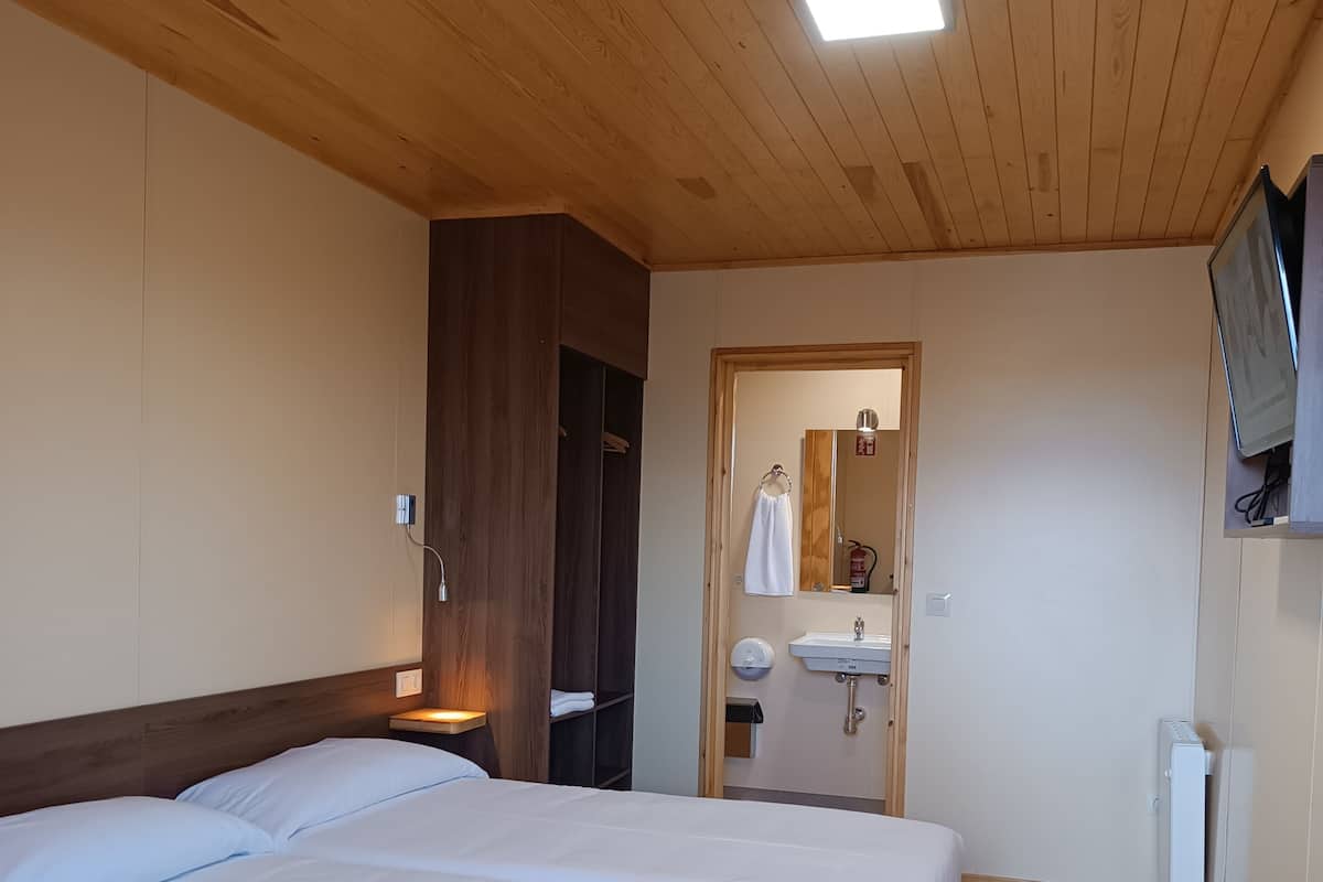A hotel room with a wooden ceiling, a large bed with white bedding, a bedside table, a wardrobe, a bathroom with a sink and mirror, and a television mounted on the wall.