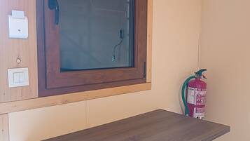 Una habitación pequeña con un escritorio de madera, una silla, un ventanal con marco de madera, un extintor y un interruptor de luz en la pared.
