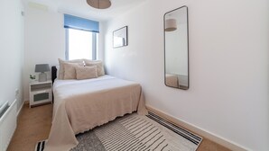 A bedroom with a bed, bedside table, lamp, window, and a mirror. - The Stratford Apartments (London)