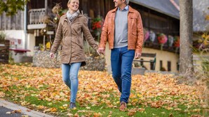 Un couple se promène sur un sentier recouvert de feuilles dans un cadre rural, avec un bâtiment en bois en arrière-plan.