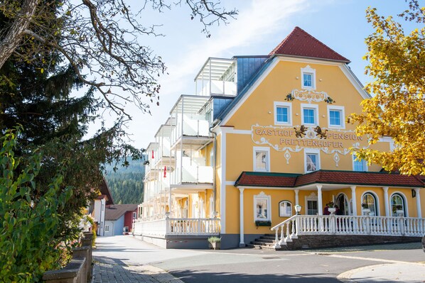 Un edifici groc amb teulada vermella, anomenat "Gasthof Pension Frettenhofer", amb un balcó i una escala que condueix a l'entrada.