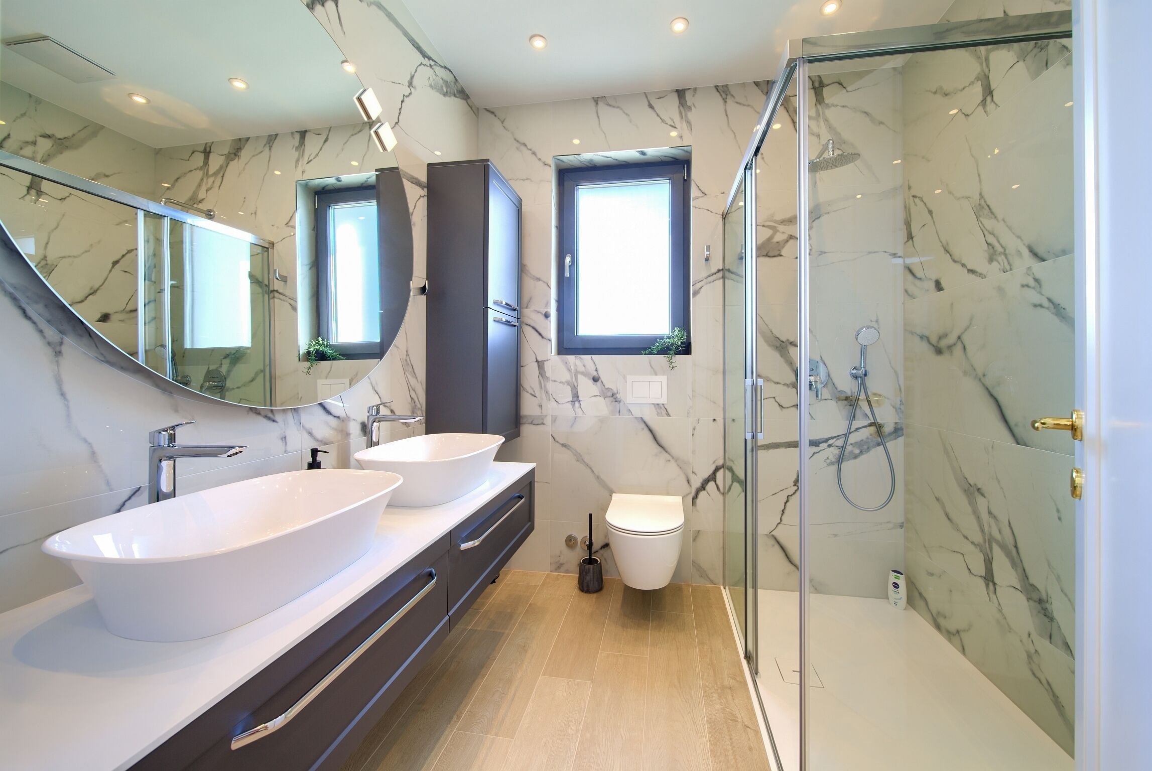 Ein modernes Badezimmer mit einem großen, weißen Waschbecken, zwei Spiegeln, einer Duschkabine aus Glas und Marmorwänden.
