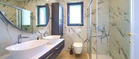 Ein modernes Badezimmer mit einem großen, weißen Waschbecken, zwei Spiegeln, einer Duschkabine aus Glas und Marmorwänden.