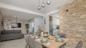 Un'area pranzo moderna con un tavolo in legno apparecchiato per sei persone, circondato da sedie. Si intravede sullo sfondo un muro in pietra e una cucina con mobili bianchi.