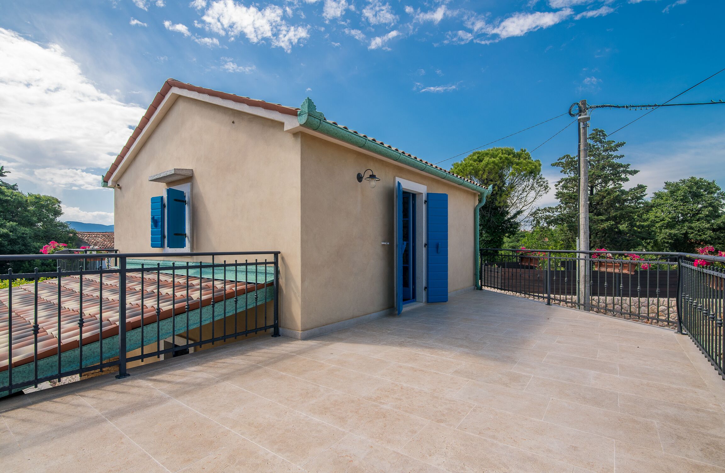 A small, single-story building with a beige facade, blue shutters, and a tiled patio area.