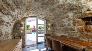 Una habitación con paredes de piedra, una mesa y sillas de madera, un vidrio que da al exterior, y un paraguas rojo afuera.