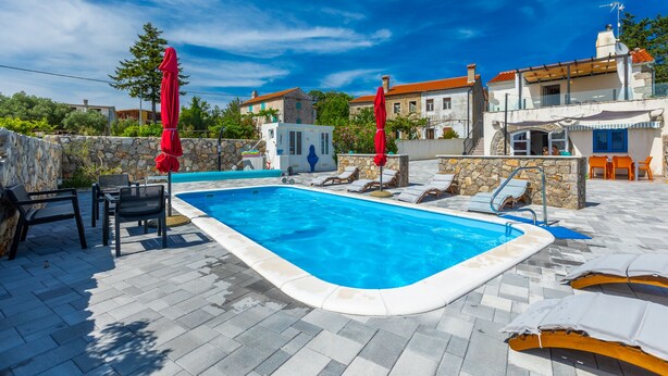 Una piscina con sillones de descanso y sombrillas, rodeada de muros de piedra y edificios con tejados rojos.