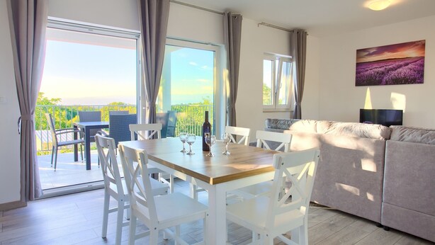 A dining area with a wooden table and white chairs, a sofa, and a balcony with a view of greenery. - Villa Elena (Kvarner - Poljica) (Poljica)