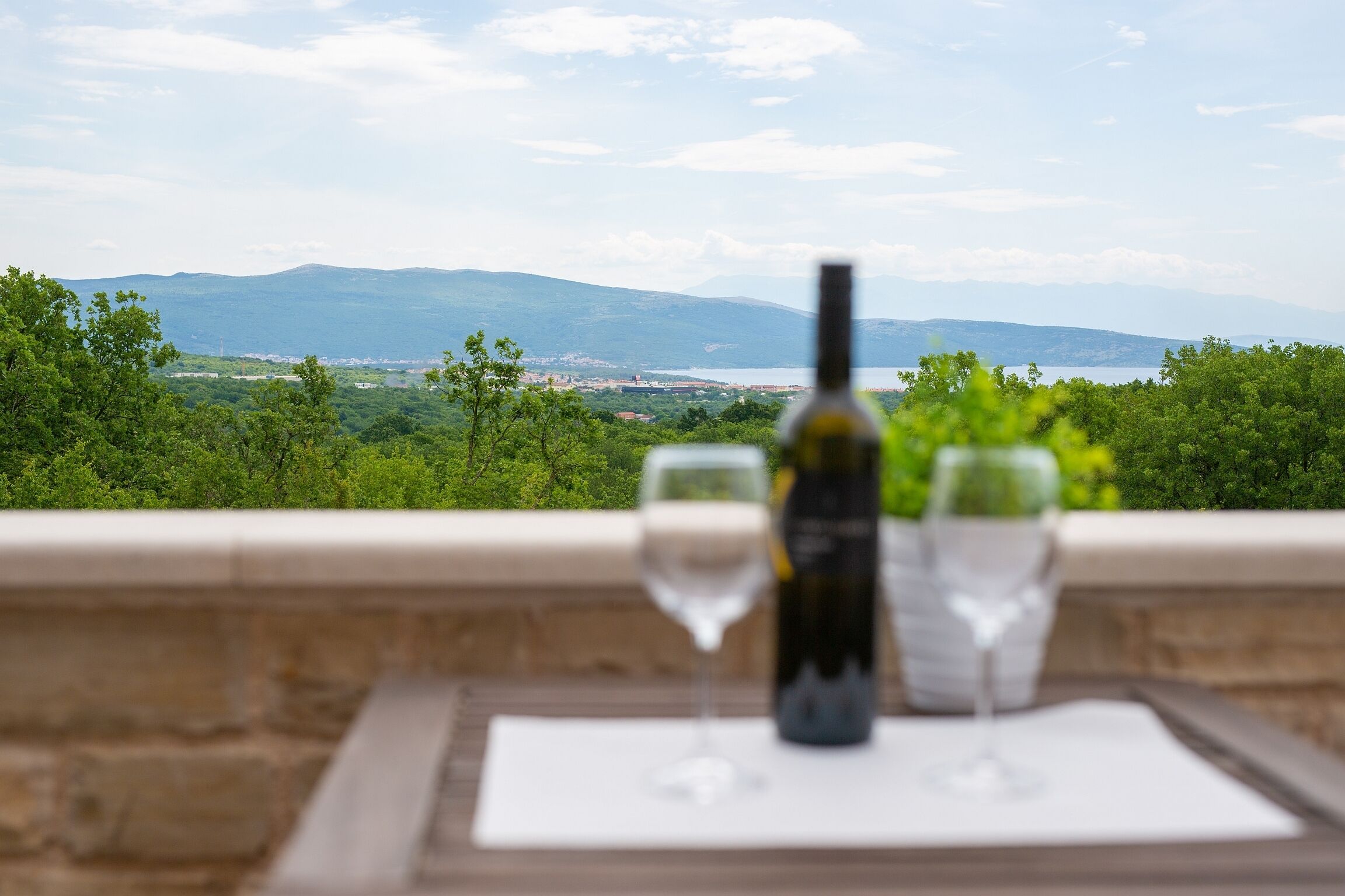 A table with a bottle of wine and two glasses on a balcony overlooking a scenic landscape with mountains and greenery.