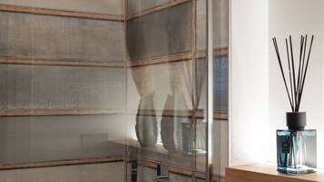 A modern bathroom with a glass partition, a wooden shelf with a bottle of reed diffuser, and a shower with a metal hose.