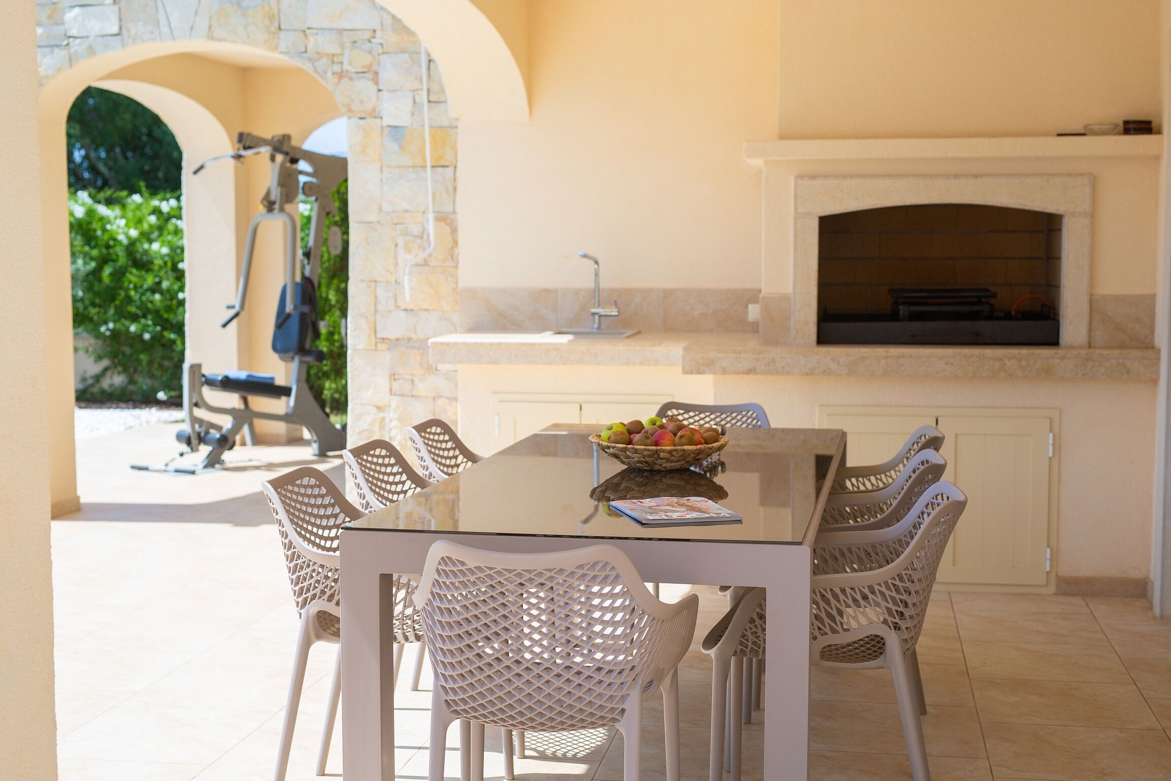 An outdoor dining area with a glass table and wicker chairs, a stone archway, and a stone wall.