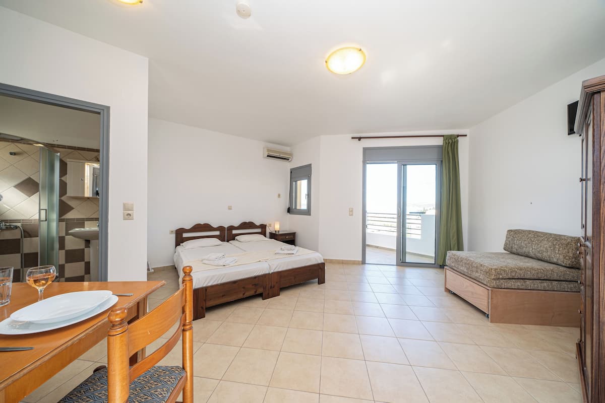 A bedroom with a bed, a bedside table, a chair, a sofa, a dining table with plates and a glass, and a bathroom visible through an open door.