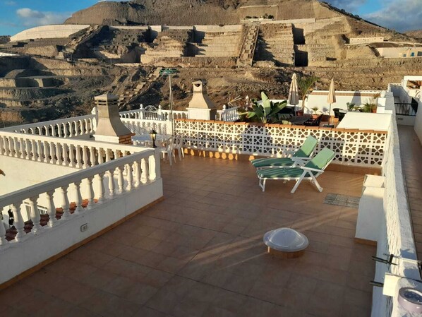 Eine Dachterrasse mit Blick auf eine gebirgige Landschaft, mit weißer Brüstung, grünen Liegestühlen und einem kleinen Tisch.