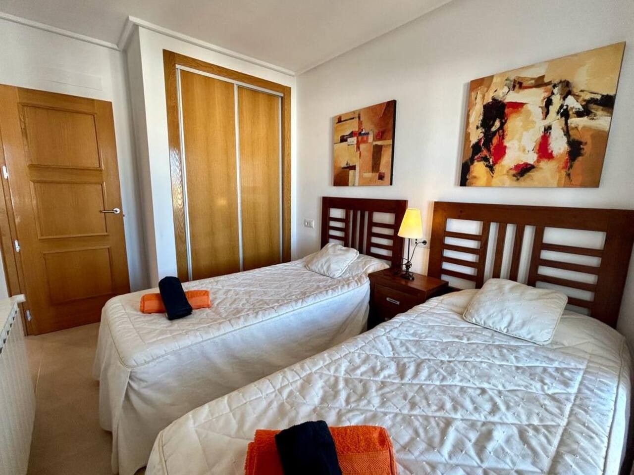 Ein Zimmer mit zwei Einzelbetten, einem Holzschrank, einem Gemälde an der Wand und einer Lampe auf dem Nachttisch.
