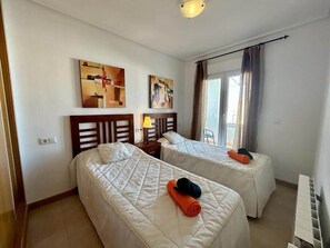 Two single beds with quilted mattresses and neatly folded towels, a bedside table with a lamp, and a wall-mounted shelf.