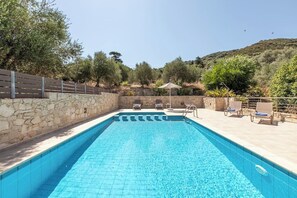 A swimming pool with a stone wall and a wooden fence, surrounded by trees and a hilly landscape.