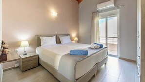 A hotel room with a bed, bedside lamps, a nightstand, a window with curtains, and an air conditioning unit. - Neaira Villas - Villa no2 with private pool (Platanias)
