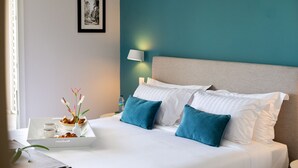 Una cama bien hecha con almohadas blancas y color turquesa, una mesita de noche con una bandeja de alimentos para el desayuno y una lámpara fijada a la pared.