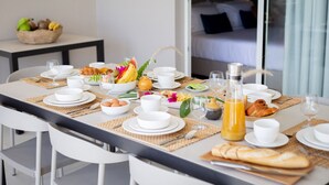 Mesa de desayuno dispuesta con platos, tazones y vasos blancos, frutas, bollería y una botella de jugo de naranja.