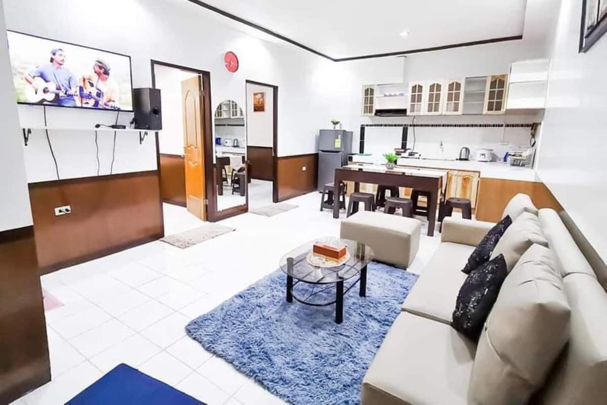 A modern living room with a sofa, coffee table, and a TV. There is a kitchen area with cabinets and a dining table.