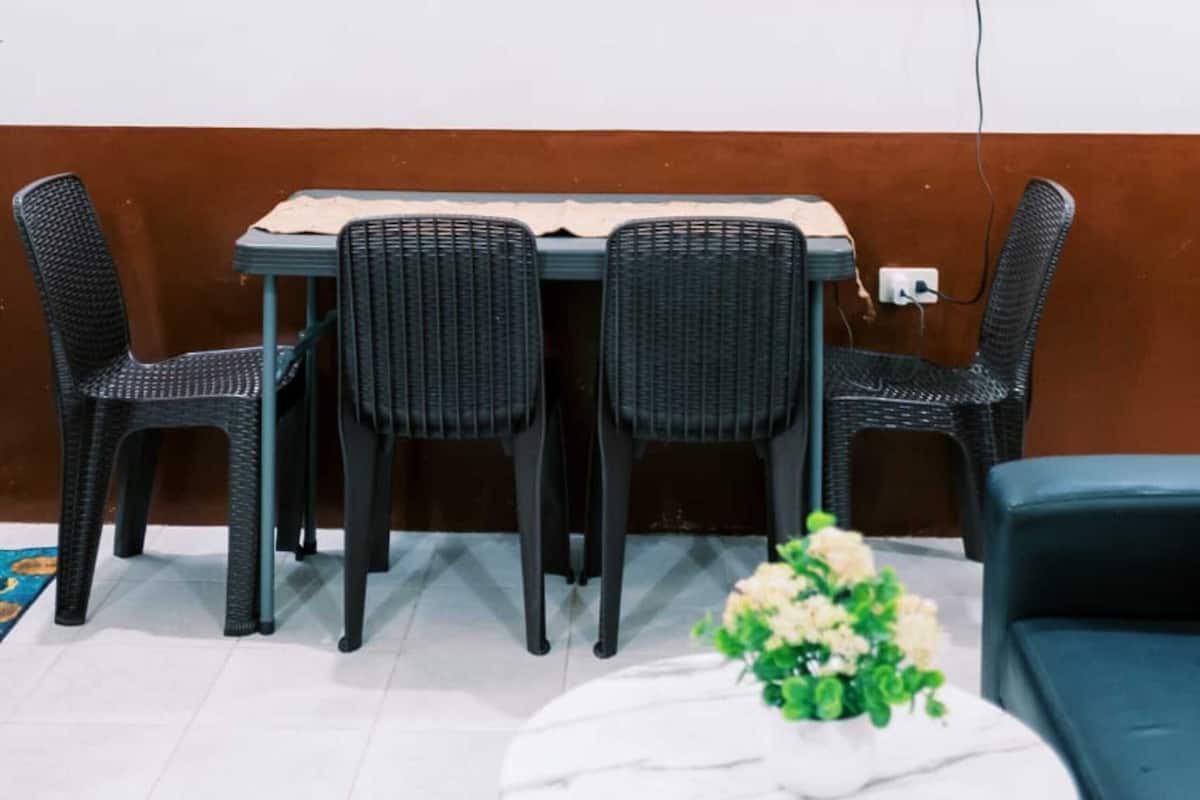 A room with a dining table, four chairs, a TV mounted on the wall, a small shelf, and a round coffee table with a flower vase.