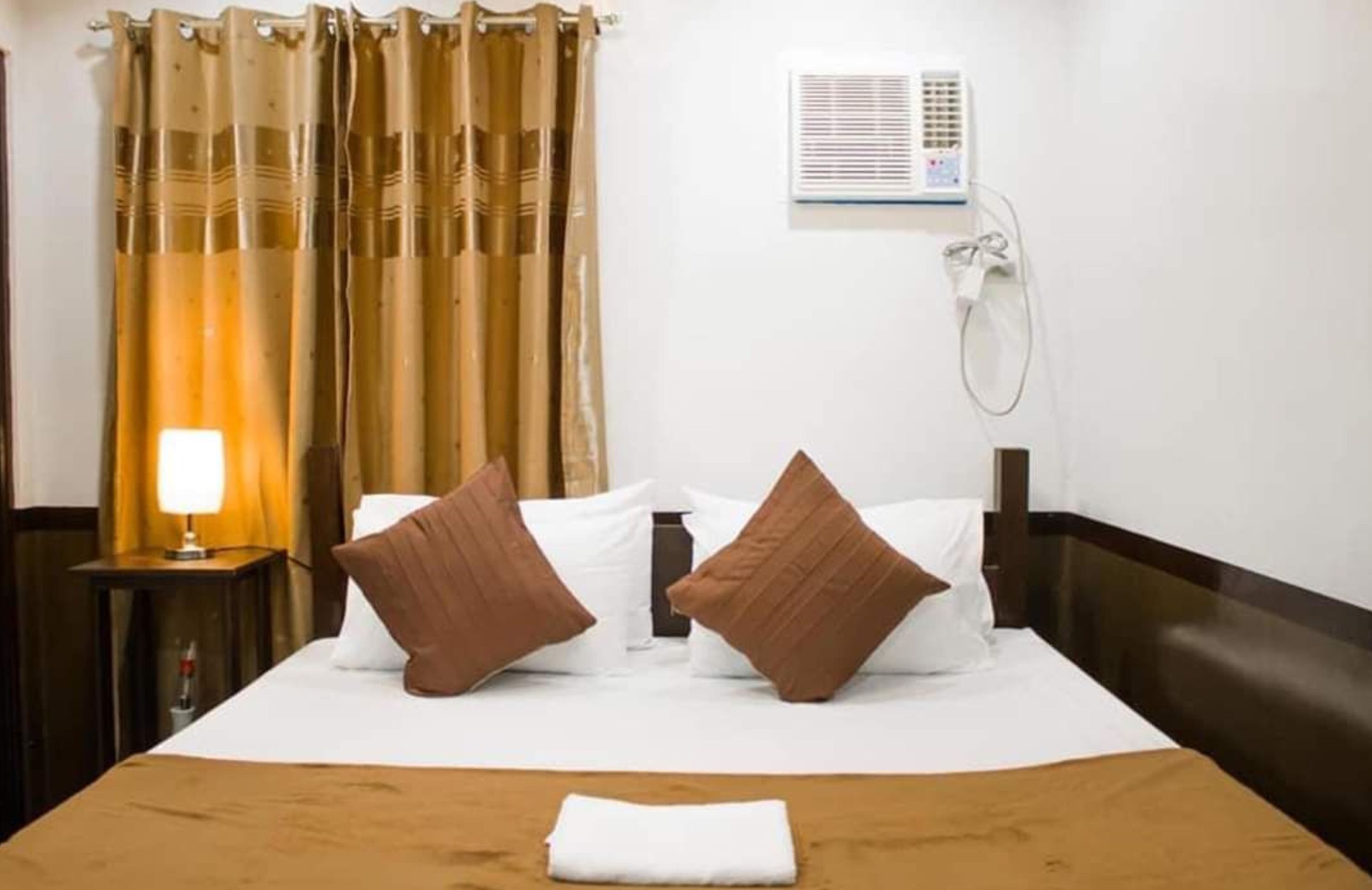 Ein Hotelzimmer mit einem Bett, braunen Kissen, einer Nachttischlampe, einem Fenster mit Vorhängen und einer Klimaanlage.