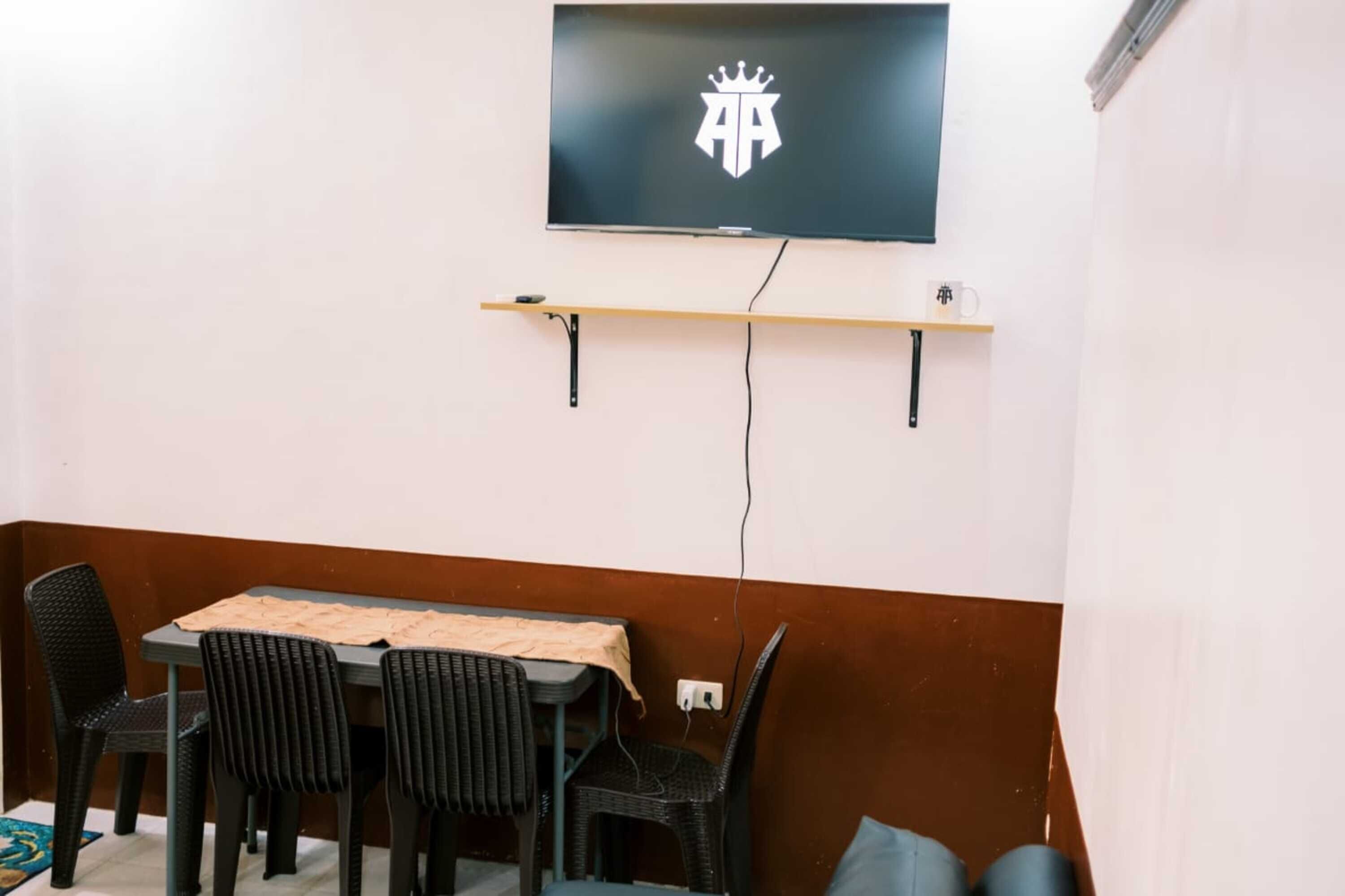 A room with a TV mounted on the wall, a small shelf below it, and a table with four chairs.