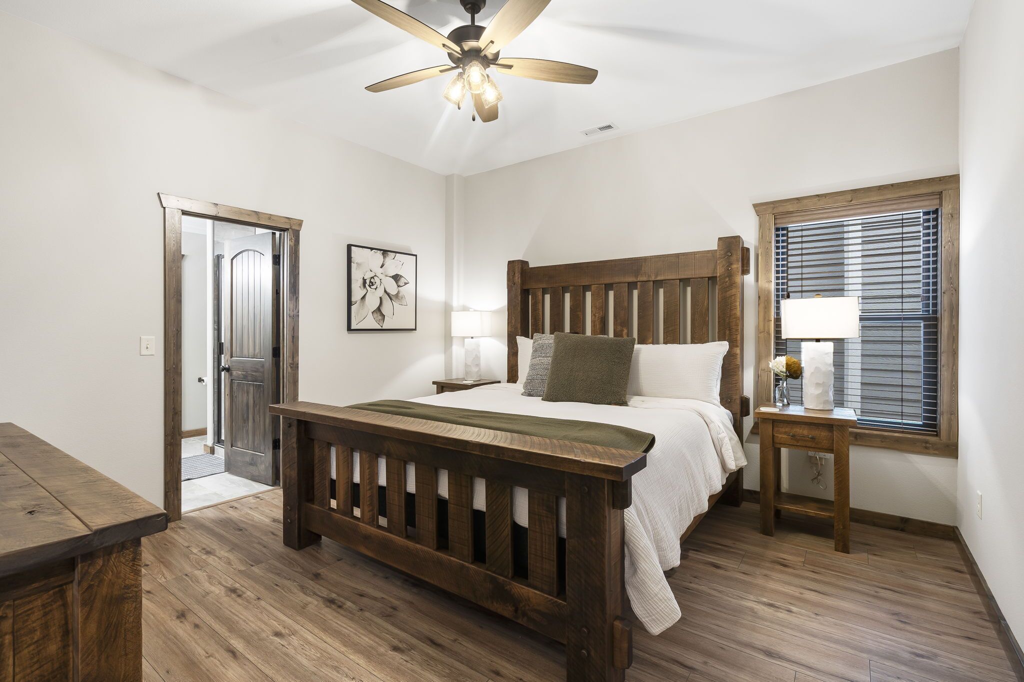 Une chambre avec un lit en bois, un ventilateur de plafond, une fenêtre avec des stores et une porte donnant sur une autre pièce.