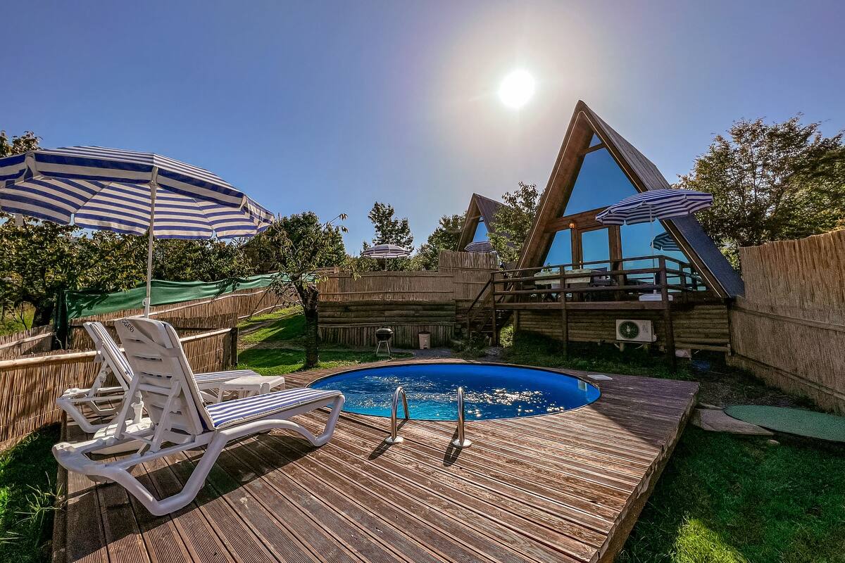 Una terraza de madera con piscina, sillones de descanso y una estructura de madera con techo triangular.