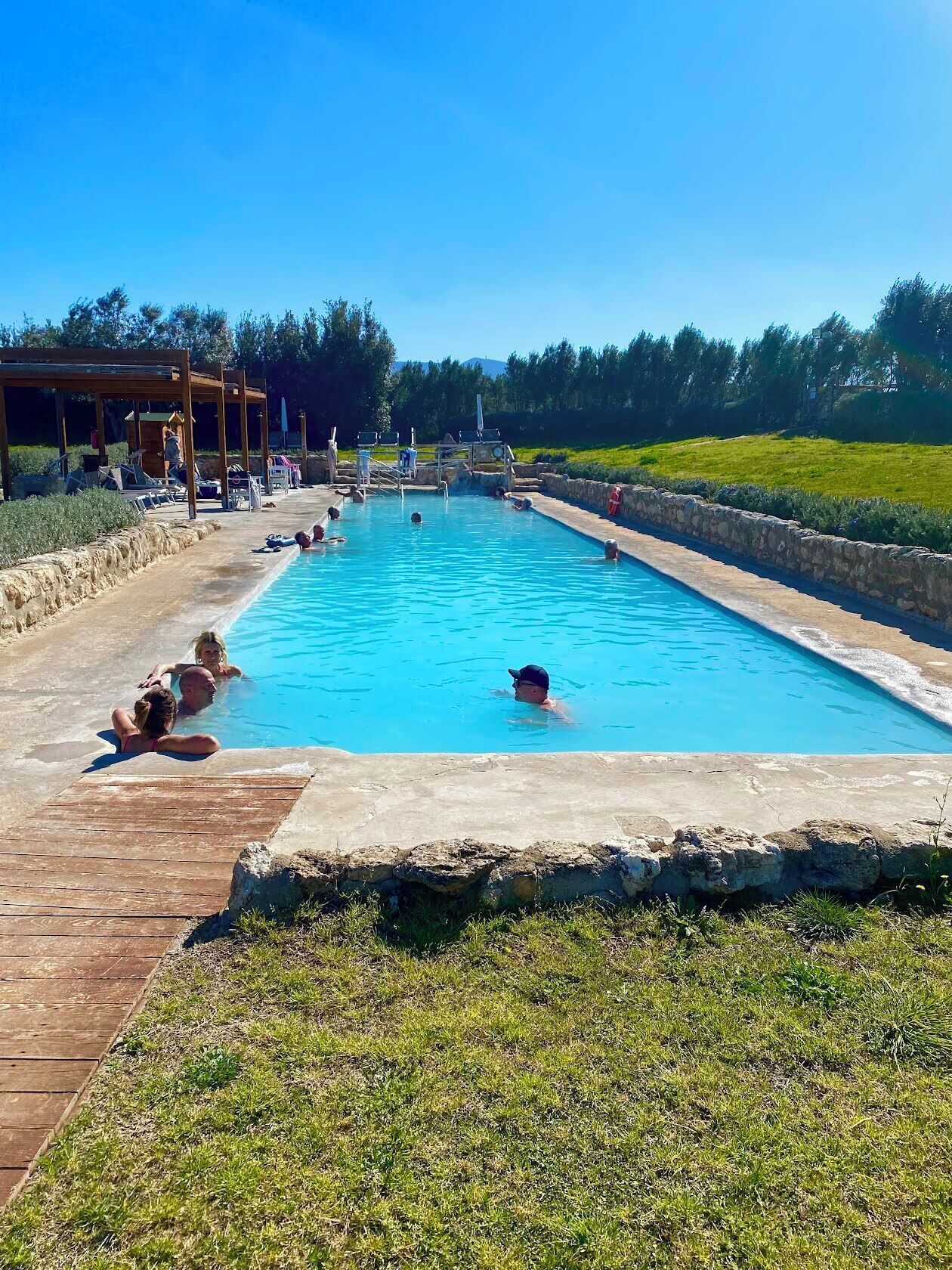 Piscina com deck de madeira e espreguiçadeiras, cercada por um muro de pedra e área gramada.
