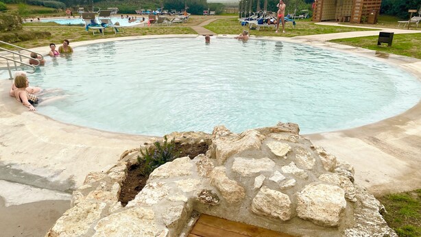 Une grande piscine extérieure, avec une structure en pierre en premier plan, entourée de chaises longues et d’une pelouse.