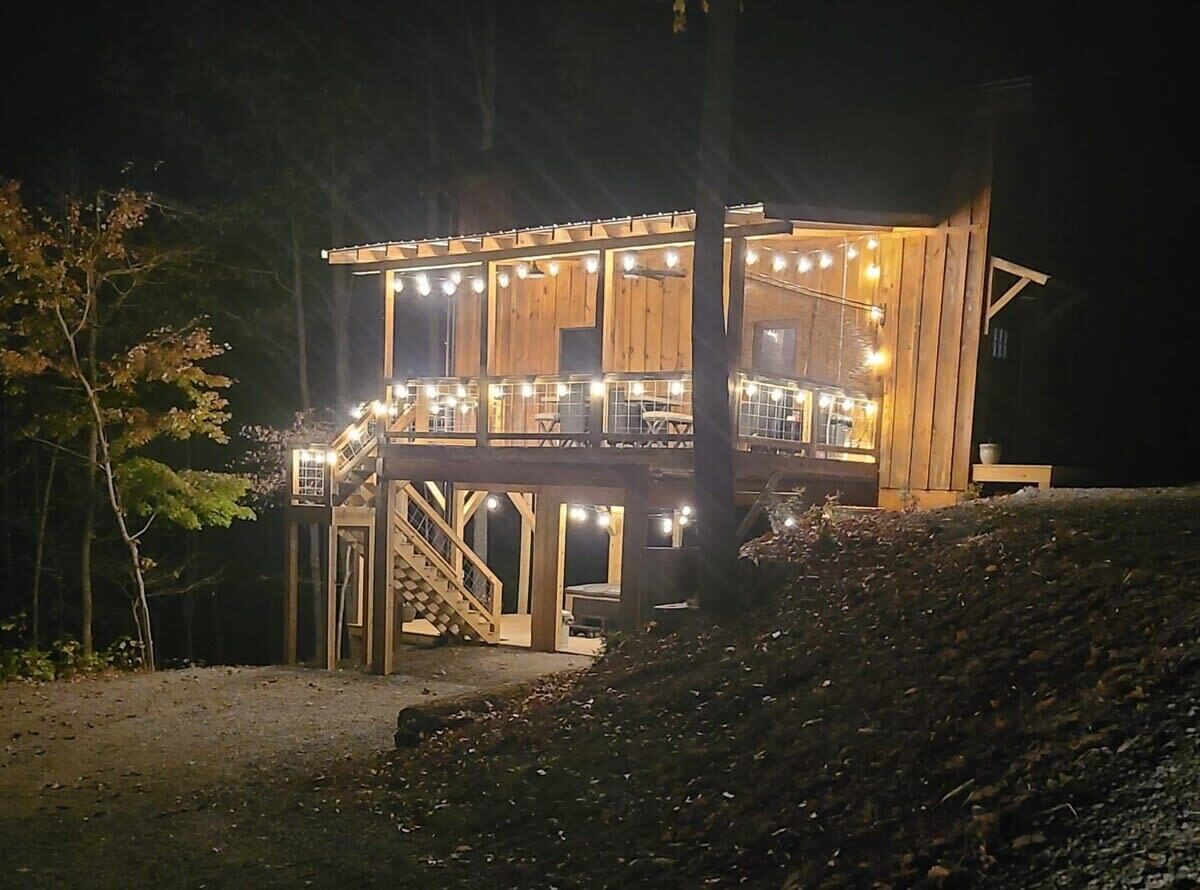 A two-story wooden cabin with a balcony, illuminated by string lights.
