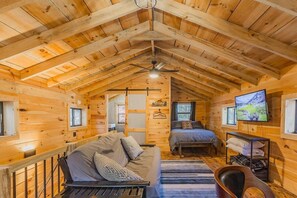 Interior acogedor de una cabaña con cama en entrepiso, sofá, televisor y comedor.