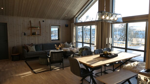 A cozy living room with a wooden ceiling, large windows, a sofa, a dining table, and a fireplace. - New mountain cabin with panoramic view, sauna, ski-in -ski-out (Innlandet)