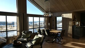 A spacious living area with a large window overlooking a snowy landscape, a wooden dining table, and a cozy seating arrangement. - New mountain cabin with panoramic view, sauna, ski-in -ski-out (Innlandet)