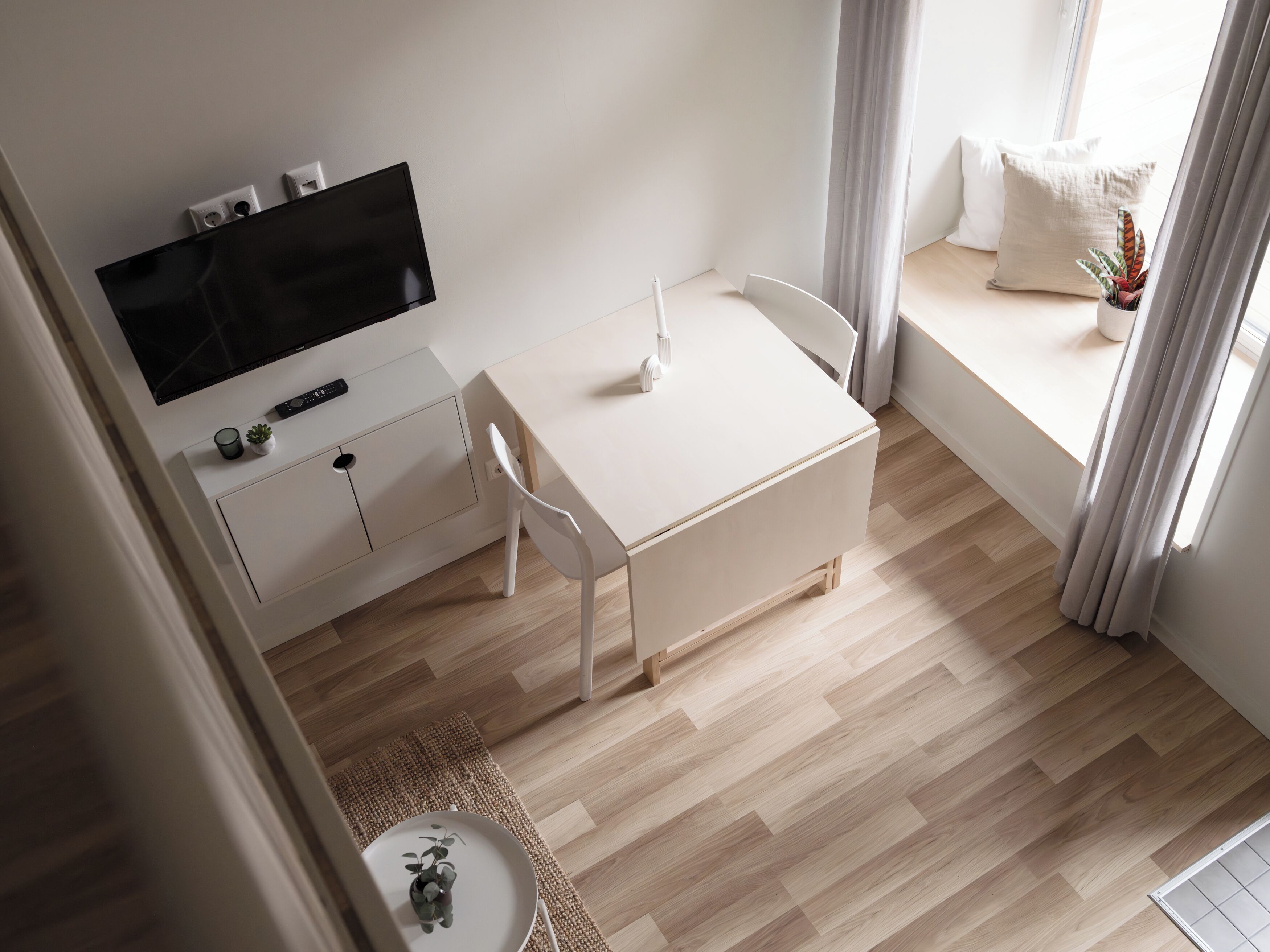 A modern, minimalist room with a flat-screen TV mounted on the wall, a white desk with a chair, and a small table with a plant.