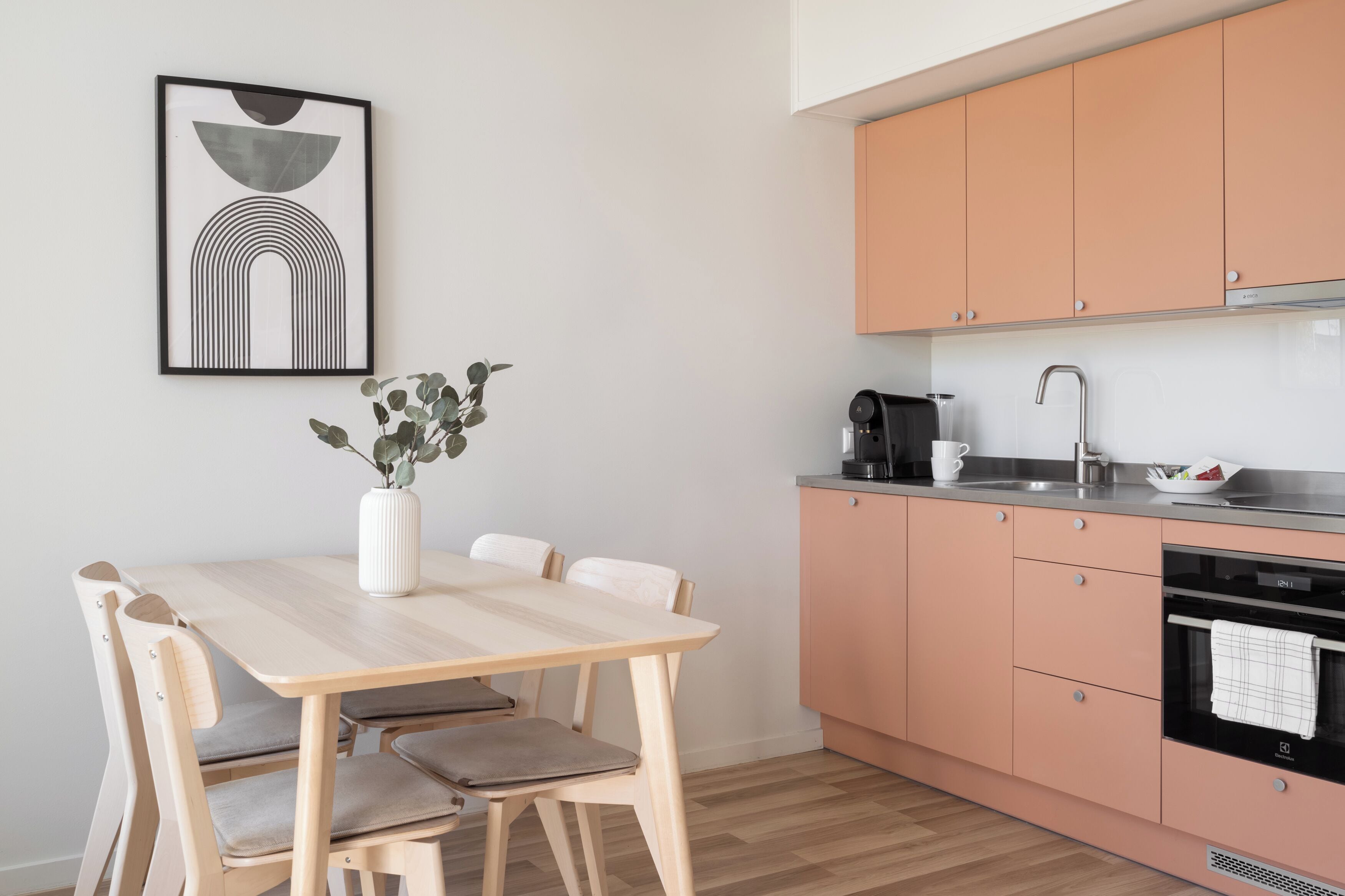 Een moderne keuken met perzikkleurige kastjes, een houten eettafel met stoelen en een lijstwerk abstract kunstwerk aan de muur.