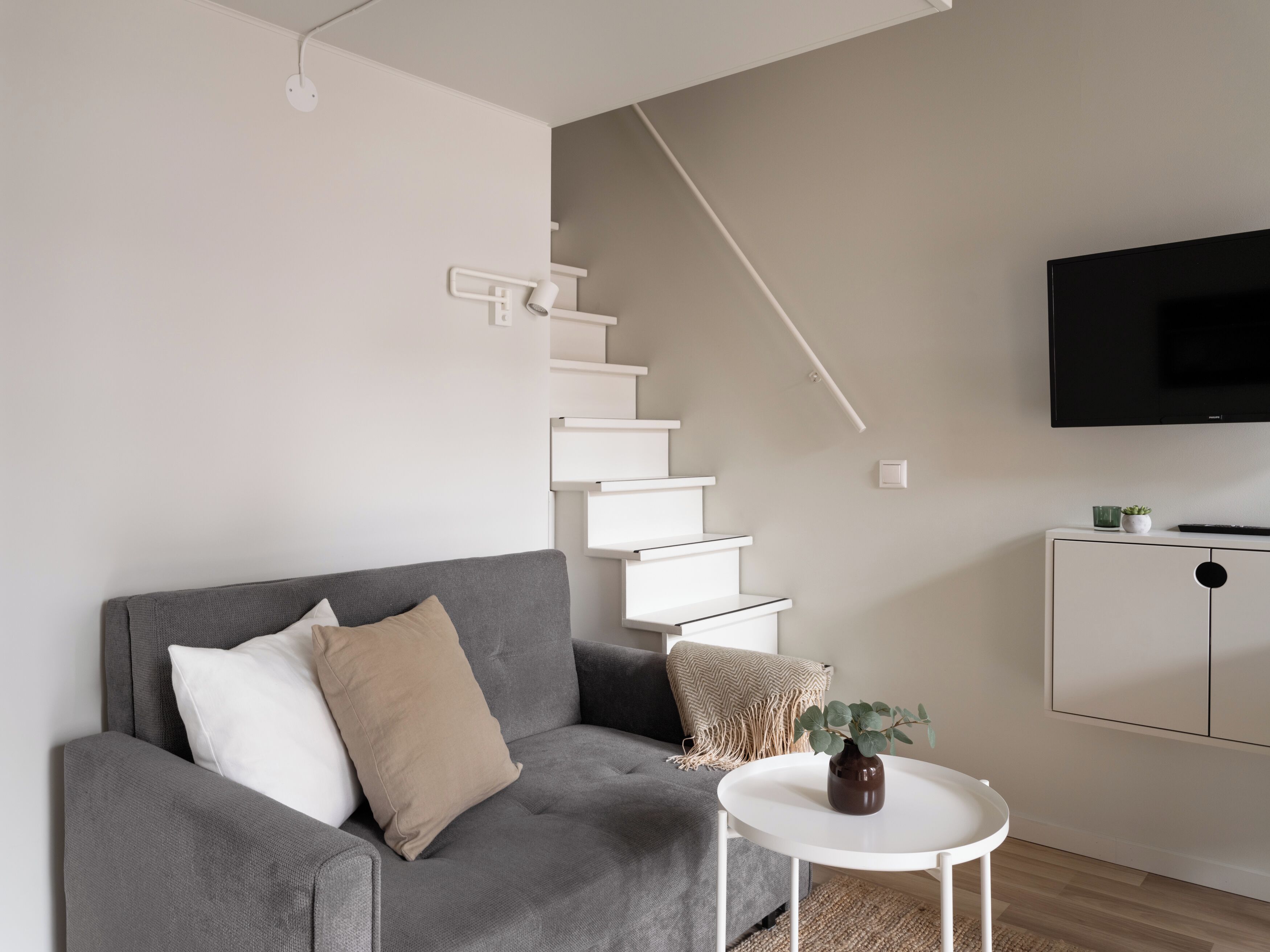 Ett modernt vardagsrum med en grå soffa, ett litet vitt bord med en vas, en trappa och en tv som är monterad på väggen.