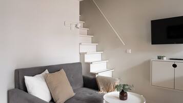 A modern living room with a grey sofa, a small white table with a vase, a staircase, and a TV mounted on the wall.