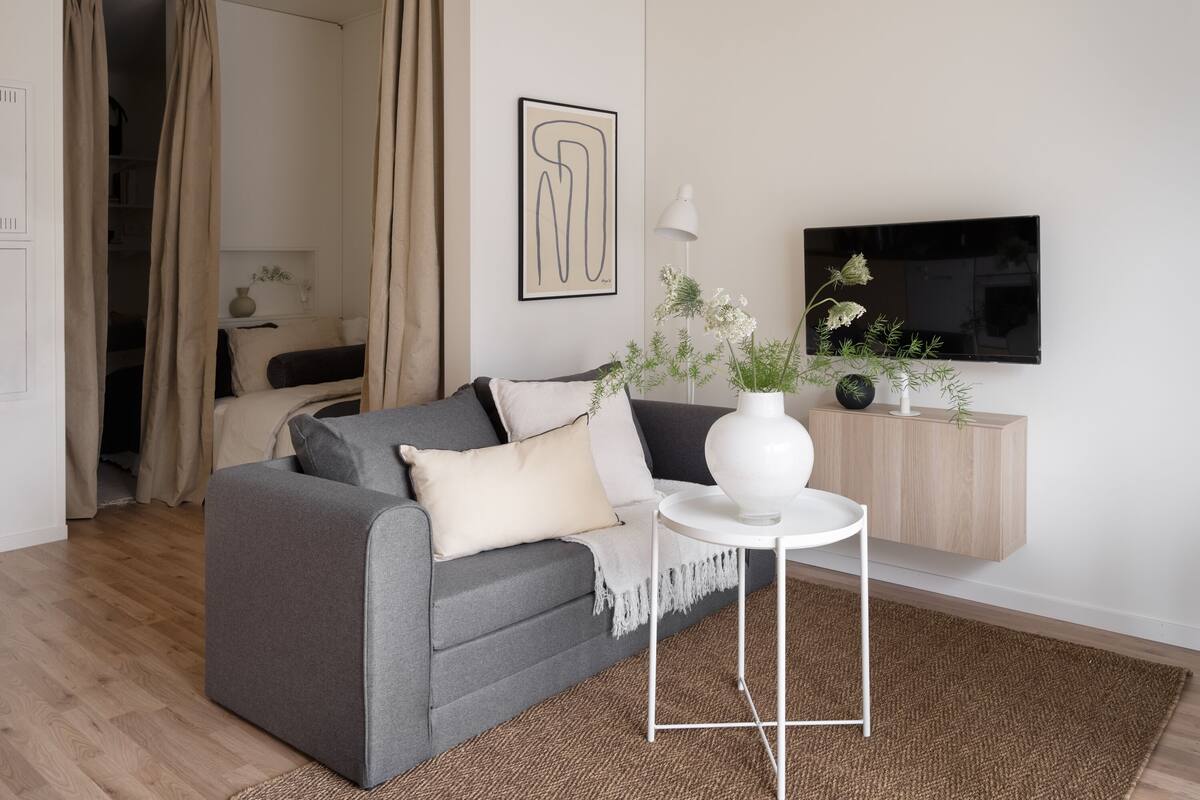 Una sala de estar moderna con un sofá gris, una mesita auxiliar blanca, un jarrón blanco con flores, un televisor de pantalla plana y un mueble de madera.