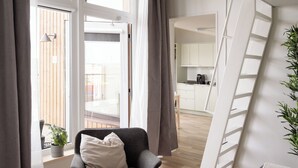 A modern living room with a large window, a grey armchair, a potted plant, and a view of a kitchen and balcony. - Waterfront Cabins Studio Apartments (Gothenburg)