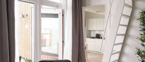 A modern living room with a large window, a grey armchair, a potted plant, and a view of a kitchen and balcony.